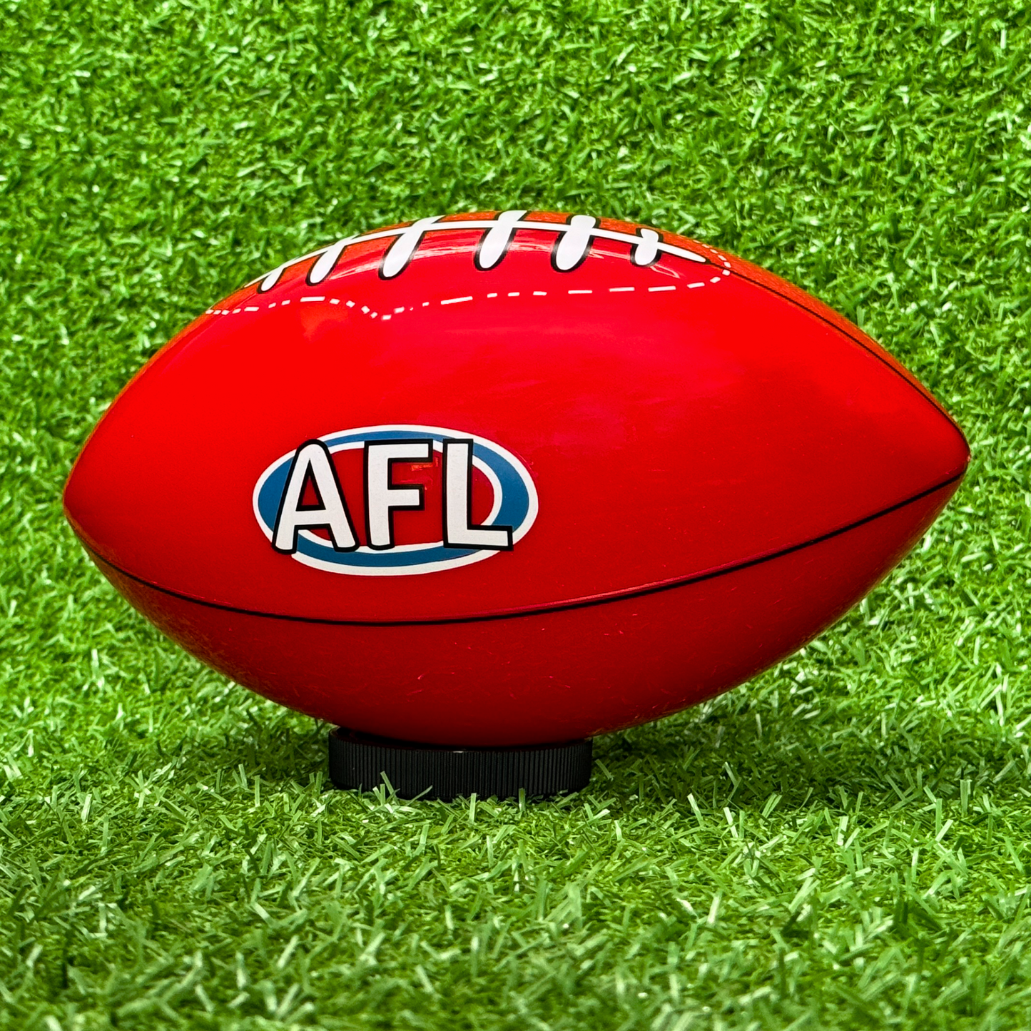 Sports Gender Reveal AFL Ball - Red Australian football with AFL logo resting on vibrant green grass, positioned on a black display stand, perfect for gender reveal celebrations and sporting announcements.