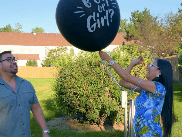 A couple outdoors playing with a giant 90CM black latex gender reveal balloon featuring "Boy or Girl?" text, set against a sunny backyard with green bushes and trees. The balloon demonstrates the product's size and outdoor party usage for Boy/Girl Giant 90CM Latex Confetti Gender Reveal celebrations.
