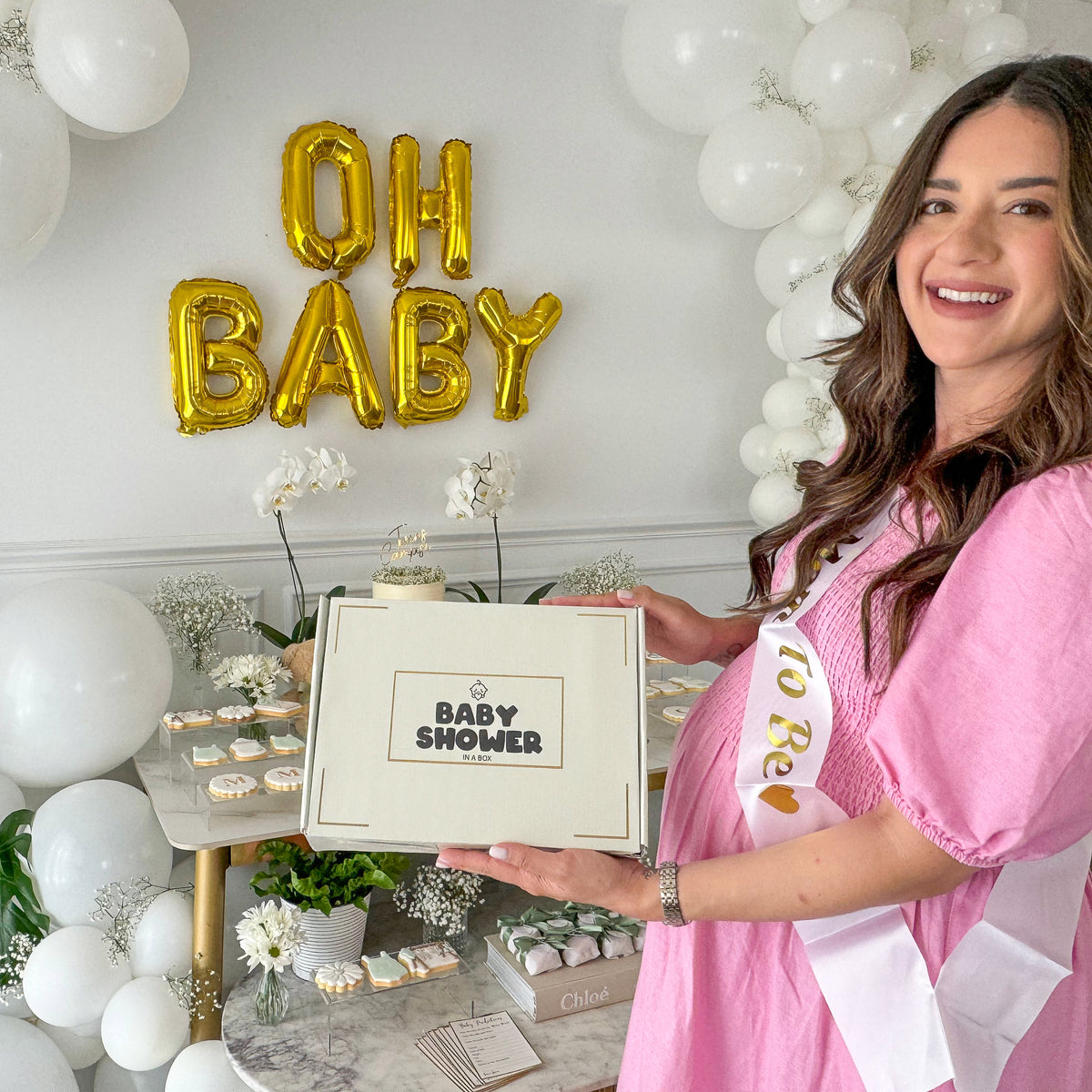 Baby Shower Party Box Set displayed at elegant celebration setup with OH BABY gold balloon letters, white balloon arch, and decorative elements. Box features neutral design perfect for baby shower hosting and party planning.